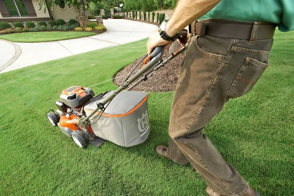A crew member in a green shirt mowing a lawn with a push mower